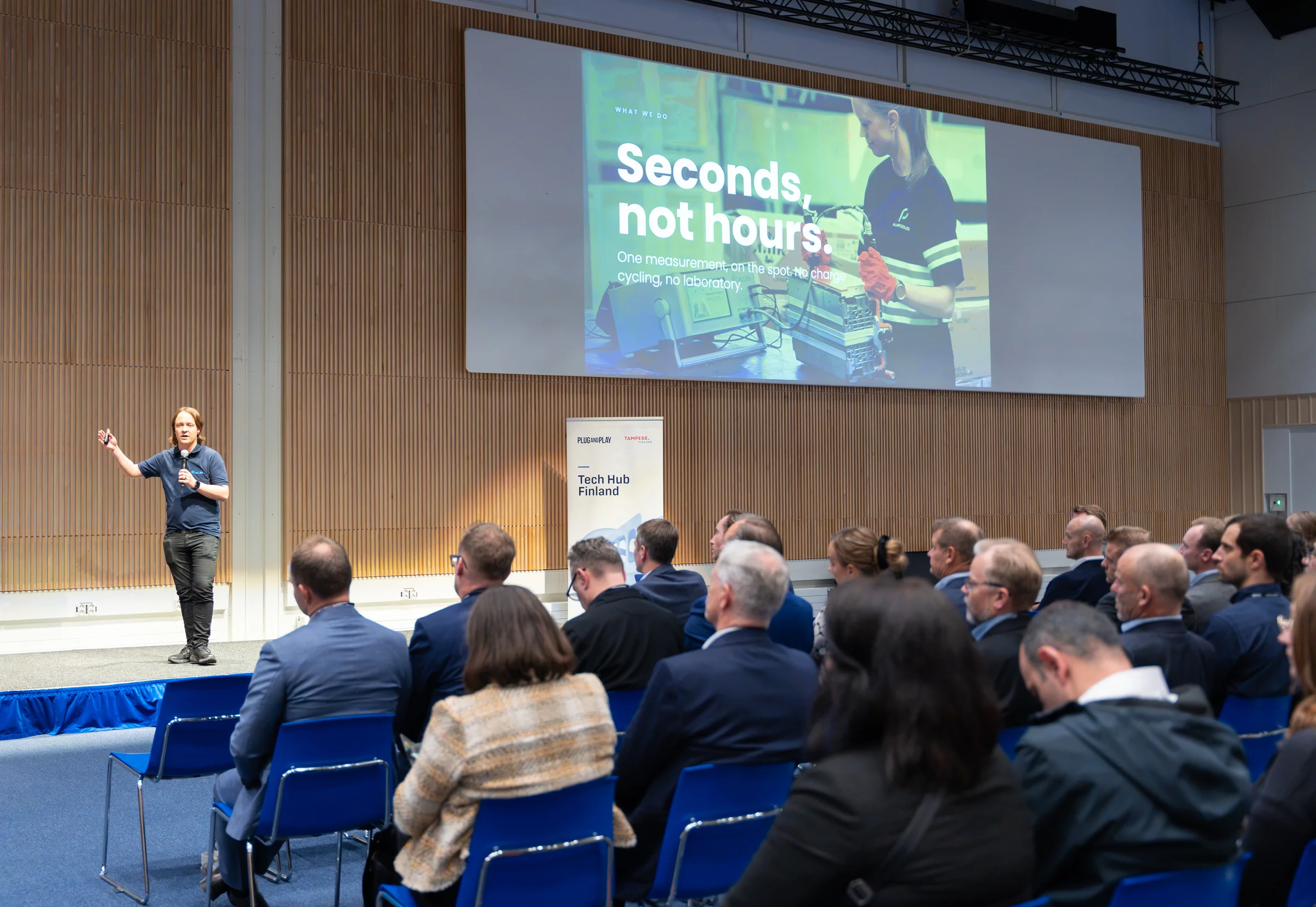 A speaker presenting to an audience beneath a large presentation screen.